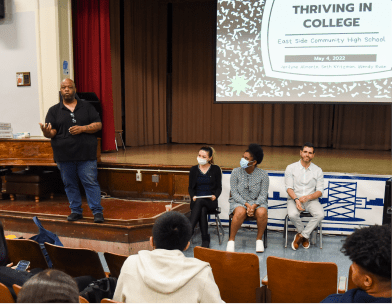 Lecturer giving a presentation titled “Thriving in College” to the students of East Side Community High School in an auditorium